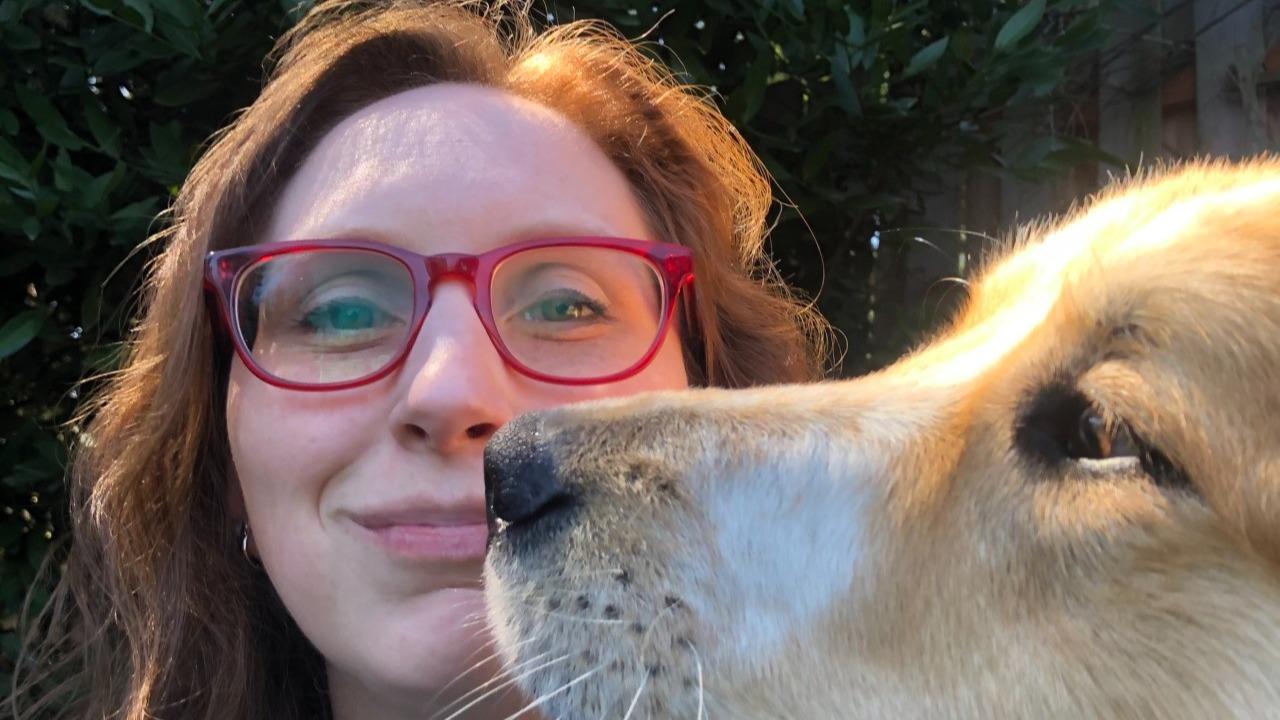 Michelle Garfinkel has a light skin tone, brown wavy hair, and wears red eyeglasses. Her dog, Lyra, is sticking her snout in the selfie frame. 