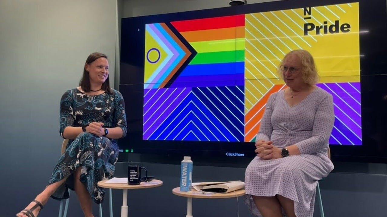 Joanne Woytek sits on a stool. Woytek has short, light-colored hair and a closed mouth smile. She wears glasses and a lavender dress. Behind Woytek is a screen displaying an LGBTQIA+ Pride Flag next to Nava's logo: a black, capital-letter N with a black line beneath it. The word Pride is beneath Nava's logo. Michelle Hadfield sits across from Woytek and smiles at her. Hadfield has shoulder length brown hair and wears a long blue patterned dress.