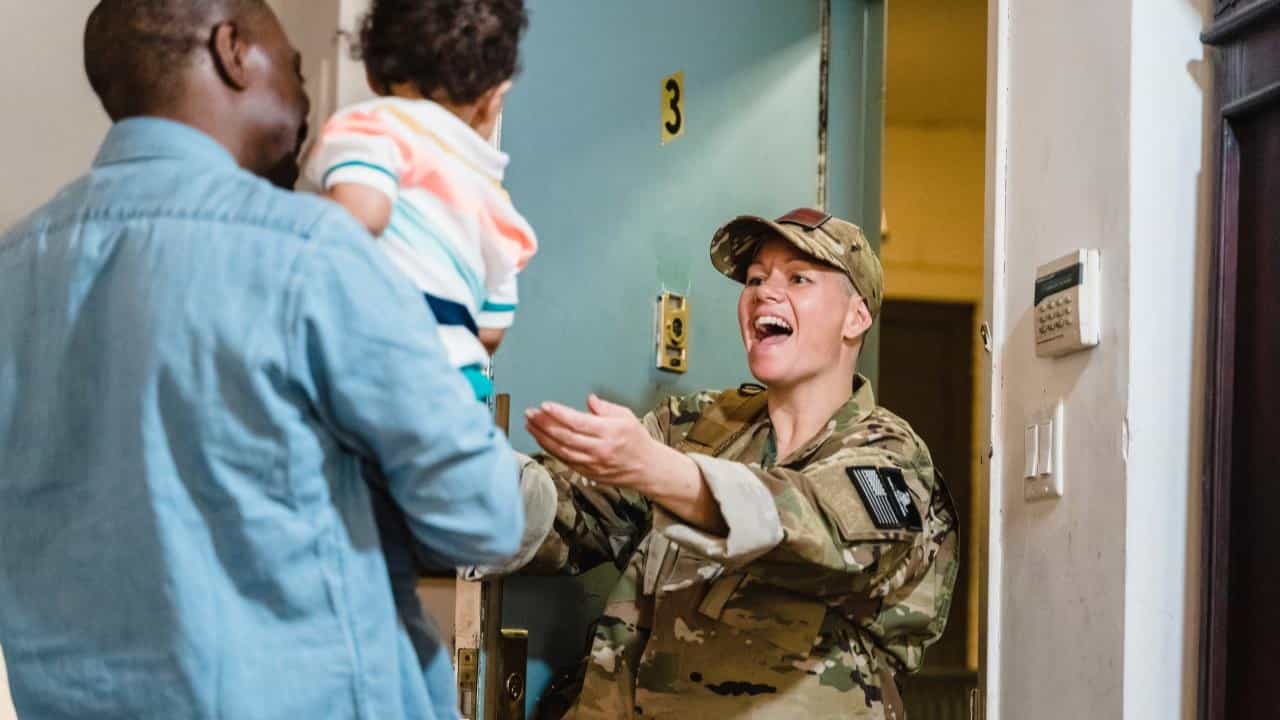 A young Black man holds a mixed-race baby and hands it to a young, white, female Veteran wearing an army uniform. 