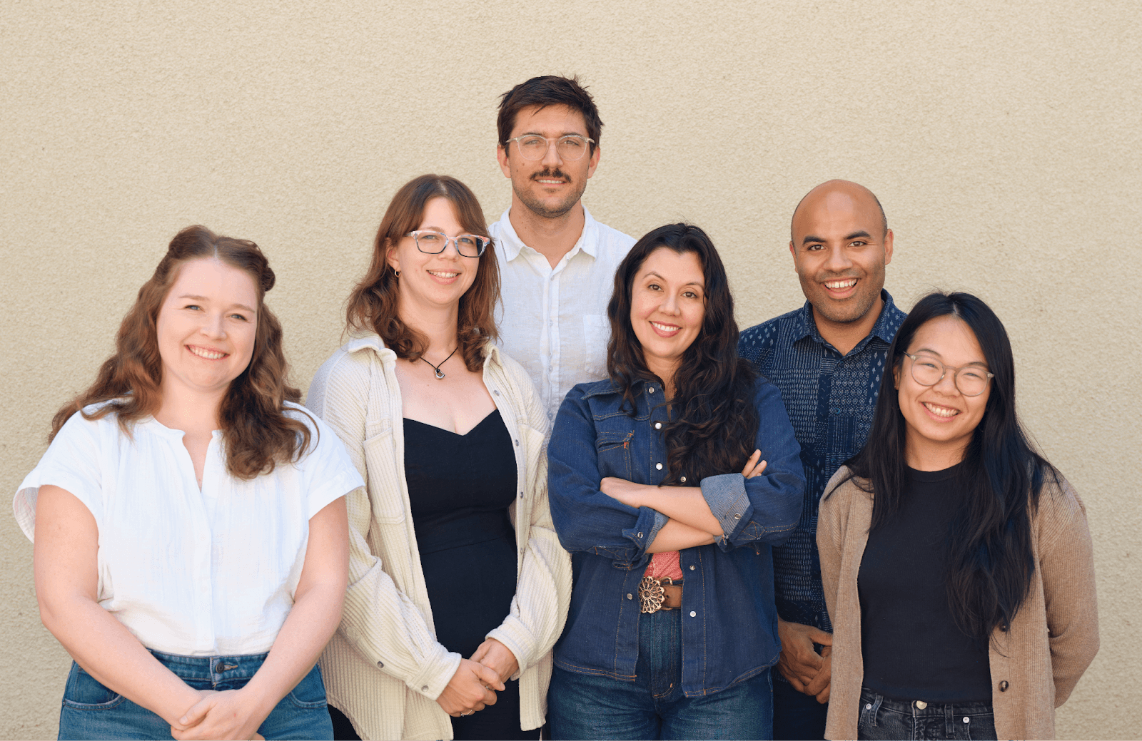 Six people stand together in front of a beige wall. From left to right: A woman with light skin and reddish-blonde curly hair wears a white blouse, a woman with light skin, brown hair and glasses wears a black shirt and cream jacket, a man with light skin, short brown hair, and a mustache wears glasses and a white collared shirt, a woman with light skin, dark brown curly hair wears a denim shirt, a man with light brown skin wears a blue collared shirt, and a woman with long black hair and glasses wears a black shirt with brown cardigan.