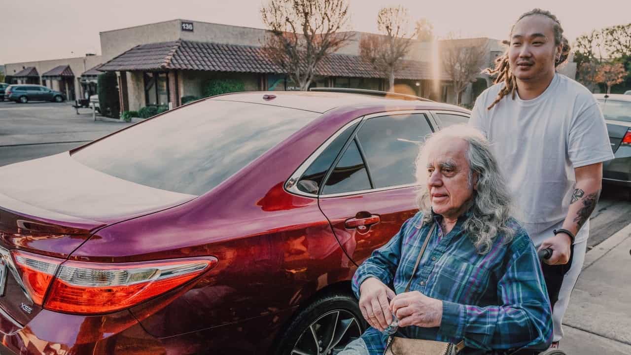 An elderly white man is pushed in a wheelchair by a young Asian man down a street sidewalk.