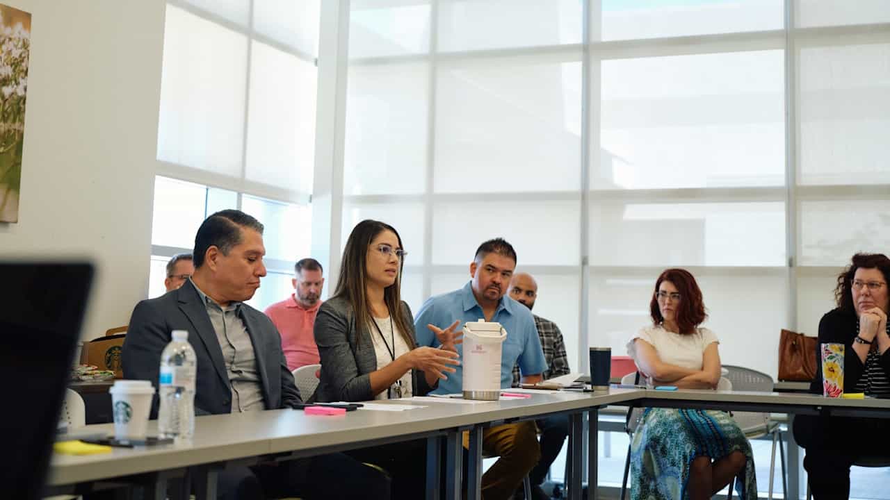 Five people sit around an L-shaped table. A woman in the center speaks and gestures with her hands.