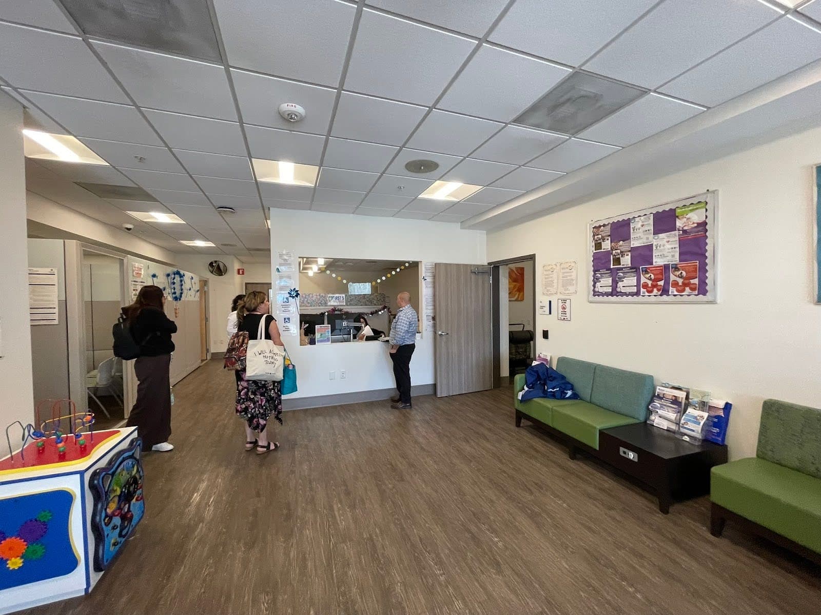 The lobby of the Family Resource Center has brown wood floors, two green sofas on the right side, children's toys on the left, and a reception desk in the center.